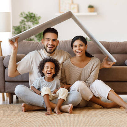 Cool-air-sfeerbeeld8-airco-warmtepomp Cheerful parents with child smiling and keeping roof mockup over heads while sitting on floor in cozy living room during relocation